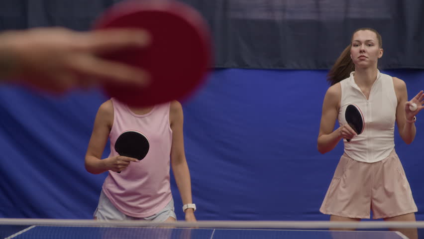 Two cheerful young women in sportswear standing on same side of tennis table rallying ball together as doubles team in competition hall - Powered by Shutterstock - Get 15% off with code: PIKWIZARD15