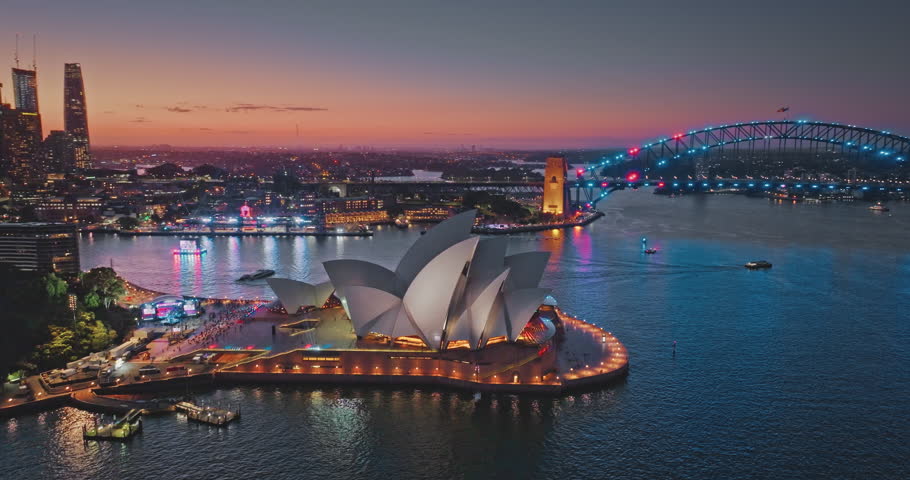 Australia, Sydney: Sydney Opera House and city skyline shining at colorful sunset, illuminated buildings and bridge creating a stunning ocean bay cityscape. Aerial view drone flight footage panorama