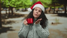 Woman wearing santa hat outdoors in park holding red cup sending kiss to camera with festive, cheerful expression against blurred trees as background during daytime - Powered by Shutterstock - Get 15% off with code: PIKWIZARD15