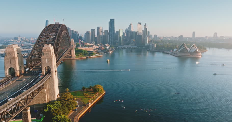 Australia, Sydney: Aerial view of Sydney Harbour Bridge, Sydney Opera House and city skyline reflecting in oean water during sunny morning, with boats sailing and cars driving. Drone flight footage