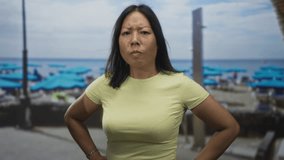 Woman clenching fist with hands on hips on a street by the beach with blue umbrellas and yellow shirt; anger defiance. - Powered by Shutterstock - Get 15% off with code: PIKWIZARD15