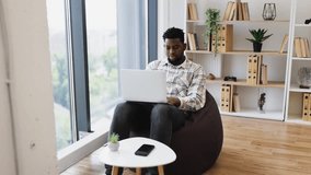Young adult man sitting on beanbag in modern home office, working on laptop, showing positive gesture. Bright setting with natural light and organized shelves in background creating calm workspace. - Powered by Shutterstock - Get 15% off with code: PIKWIZARD15