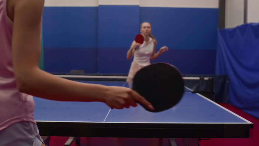 Cropped shot of concentrated young woman in sportswear returning ball to opponent over net while playing table tennis in competition hall, focus on racket