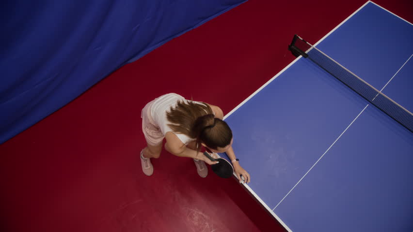 High angle shot of focused young woman in activewear tossing ball high into air then serving slice shot during table tennis match in competition hall, slow motion