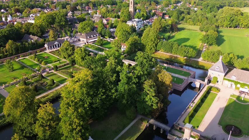 Aerial view of a formal garden with geometric layout, bordered by a canal and trees. A white manor stands at the center, surrounded by paths, hedges, and elegant landscaping.