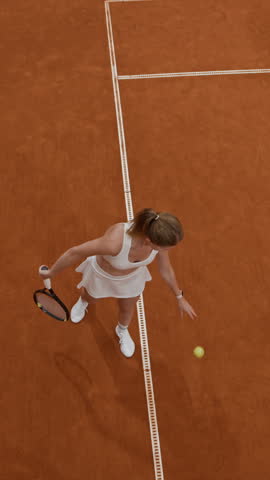 Vertical high angle shot of focused young woman in white tennis set bouncing ball against clay court then serving strong overhead shot during sports match, slow motion