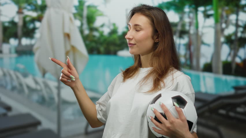 Young woman with a soccer ball points at a beautiful outdoor hotel pool, showcasing resort vibes and leisure in a tropical setting.