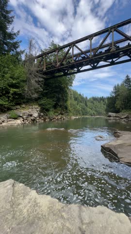 An old, rusty railway bridge spans a mountain river surrounded by a dense green forest on a bright day with a blue sky and white clouds