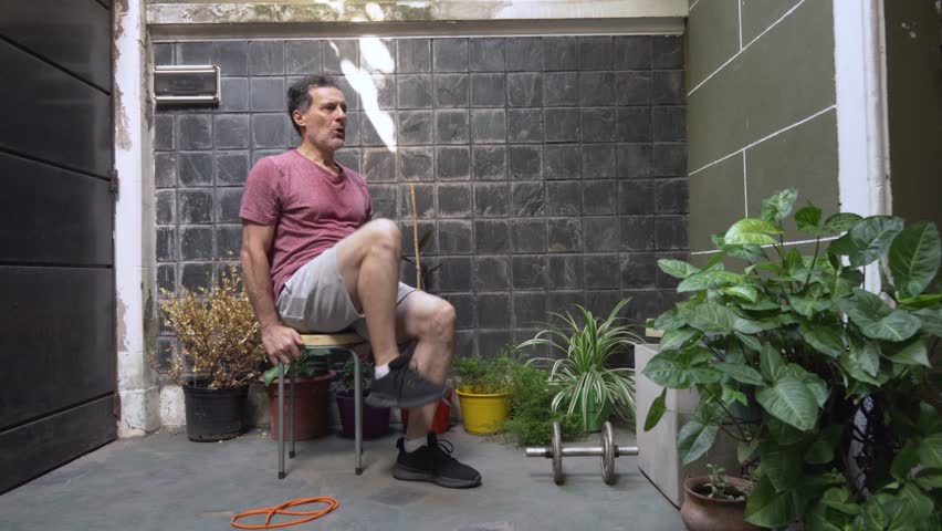 Mature man with glasses uses a chair in a domestic outdoor setting to perform a seated bicycle exercise, targeting his abdominal and oblique muscles.