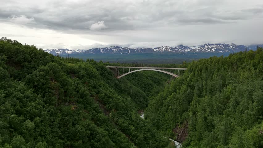 Aerial View of Van Driving Across Bridge High Above Ravine with Trees, Mountain Range with Snow in Background, lots of clouds, mid afternoon, Alaska Range, Hurricane Gulch Bridge on Parks Highway - Powered by Shutterstock - Get 15% off with code: PIKWIZARD15