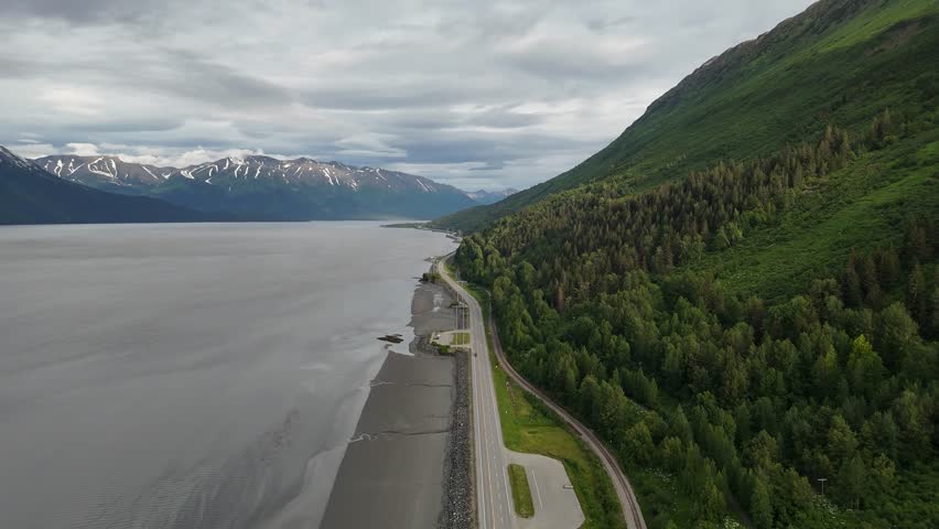 Aerial View of Seward Highway in Alaska along Turnagain Arm, Tall Green hills with trees and mountains with snow, cars driving down road, cloudy sky, Inlet of water