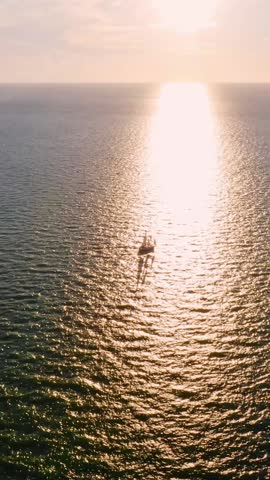 A lone sailboat glides across calm ocean waters, silhouetted against the horizon where the setting sun spreads a golden reflection across the shimmering sea.
