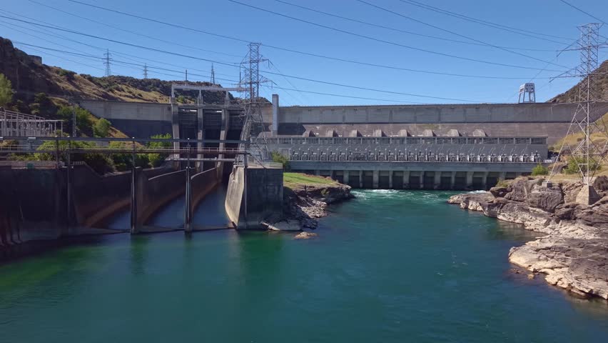 Aerial drone shot flying in reverse low over the Clutha River, revealing the full span of Roxburgh hydroelectric dam in Central Otago, New Zealand.