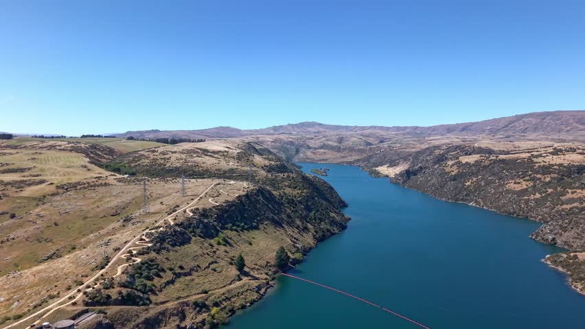 Stunning aerial drone shot flying diagonally to reveal the blue waters and surrounding hills of Lake Roxburgh in Central Otago, New Zealand.