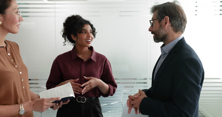 Diverse business colleagues standing together in office space, talking on work break, discussing collaboration, teamwork, sharing ideas for work on project, chatting, smiling