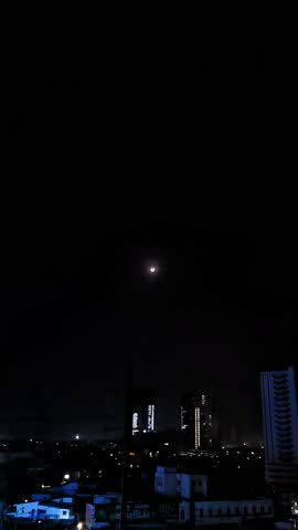 Time-lapse of a bright Moon descending over a dark city at night. Features dramatic contrast between the glowing celestial body and urban building silhouettes. 