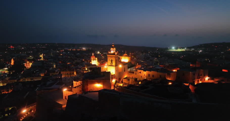 Malta, Comino island: Night lights illuminate the Citadel and surrounding buildings in the capital city of Victoria, Gozo, creating a mesmerizing cityscape aerial view. Drone flight footage