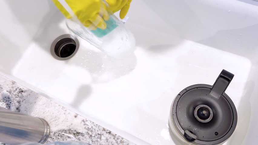 Yellow-gloved hands are seen washing dishes, scrubbing diligently in a basin sink, surrounded by suds, cleaning utensils, and clear water glasses.