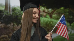 Smiling woman in graduation cap and gown holds american flag in green park; pride achievement celebration success. - Powered by Shutterstock - Get 15% off with code: PIKWIZARD15