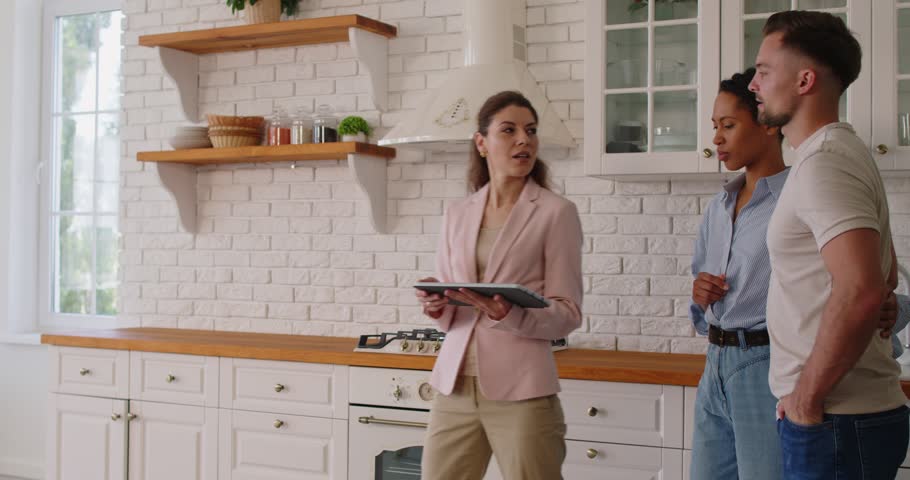 Real estate agent showing property and modern kitchen interior to couple on home apartment tour. Realtor with tablet and clients walking with discussion, broker pointing on room to man and woman