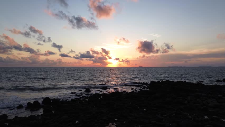Drone footage of a dramatic colorful sunset in Koh Lanta, Thailand, with rocky shoreline, calm ocean waves, and small islands across the glowing horizon in far distance.
