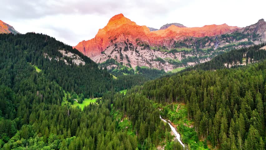 Simmenfalle waterfall tumbles through dense evergreen forest toward a sunlit alpine peak at golden hour, serene Bernese Oberland landscape, Lenk, Switzerland, drone establishing shot