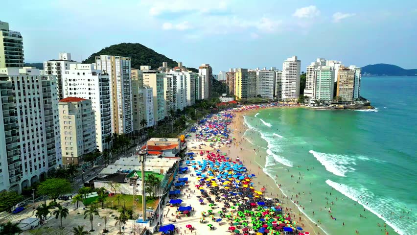 Aerial image of Guarujá beach on a very sunny day, with the beach packed with tourists under colorful umbrellas. Baixada Santista and the southern coast of the state.