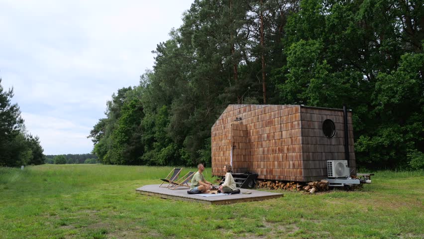 Young couple participating in a traditional tea ceremony on a wooden deck next to their modern tiny cabin, surrounded by a lush green forest and an open meadow on a peaceful summer day