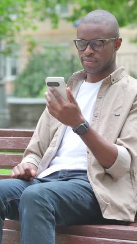 Vertical Video of Young African Man Video Calling or Conferencing on a Phone on a Park Bench
