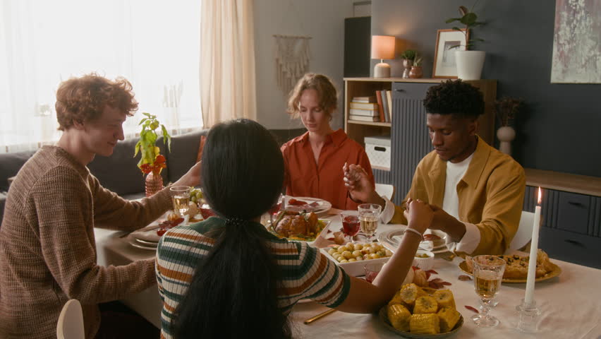 Group of multiethnic young friends finishing grateful prayer and getting down to holiday meal while sitting at beautifully set dining table in living room