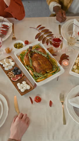 Vertical top down view of group of unrecognisable friends joining hands to say mealtime prayer before having holiday dinner at table set in fall style with baked turkey in middle