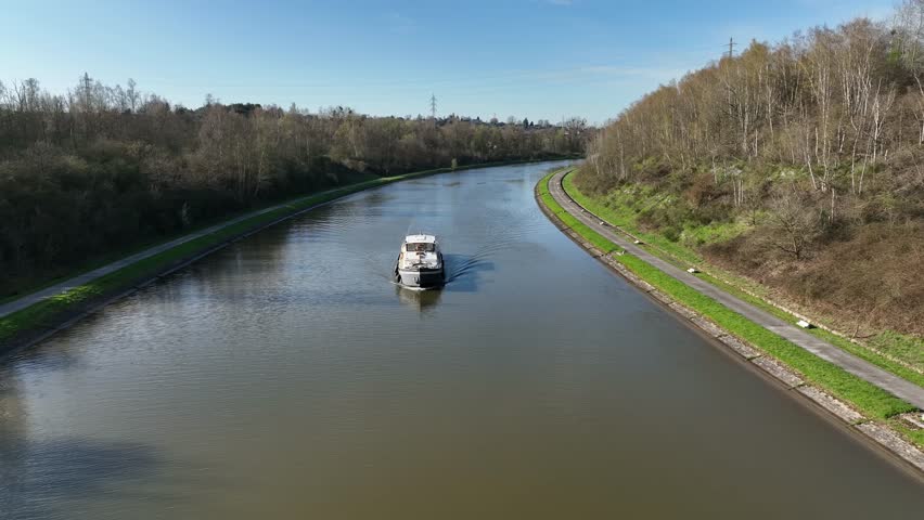 A boat advances calmly along a canal in Brussels, Belgium, surrounded by green banks and trees under clear skies.