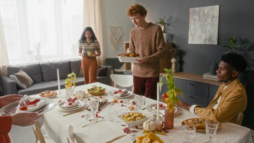 Two multiethnic girls and Caucasian young man setting holiday table with dishes and plates, African American guy sitting on chair watching