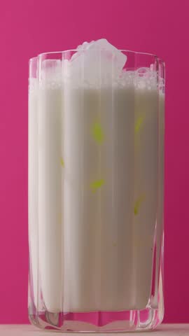 Step-by-step matcha latte preparation with milk and ice in clear glass, vibrant green tea pouring into white milk, pink background, macro.
