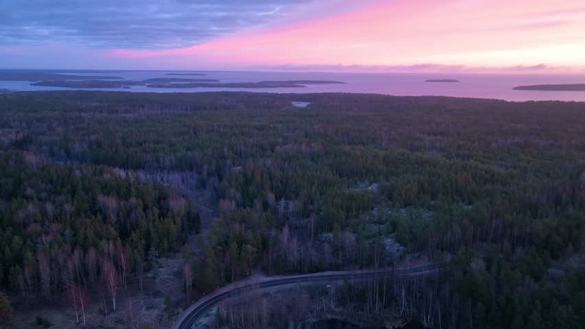 Elevated shot of a dense coniferous forest and a road, with a frozen lake and small islands in the distance under a pink and blue evening sky