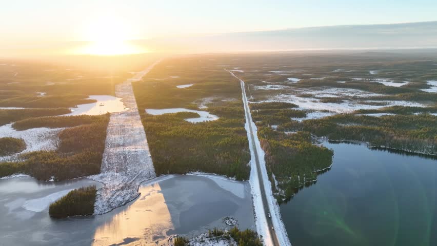 High angle shot of a straight road dividing a coniferous forest with light snow cover and a dark, calm lake, illuminated by bright sunset light