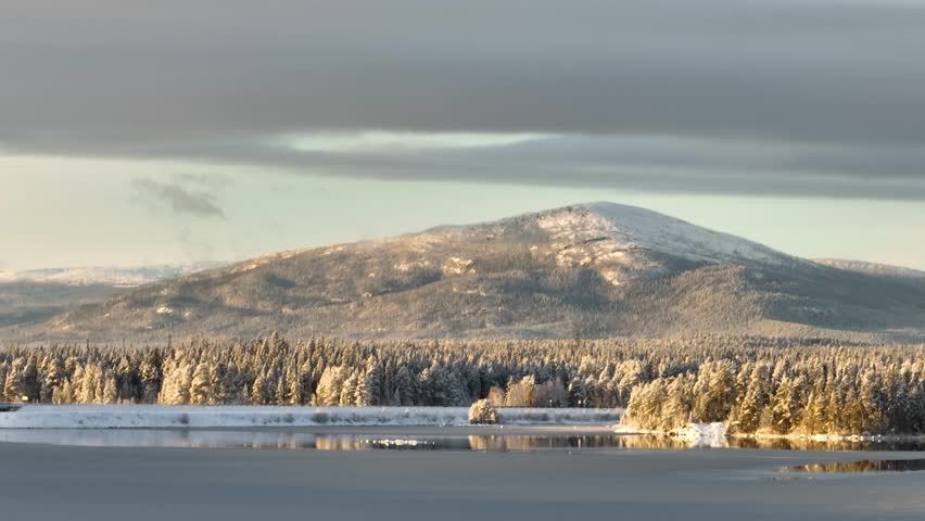 Panoramic view of a large, snow-dusted mountain rising above a dense, frost-covered forest and frozen lake under a cloudy sky during low sunlight