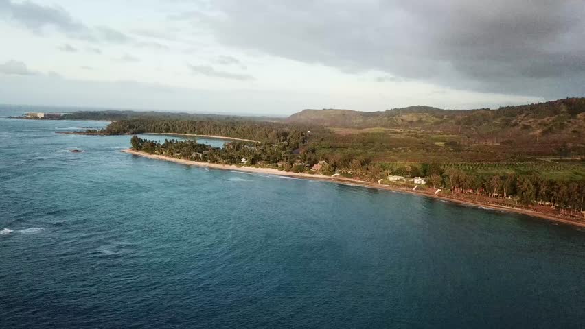 Cinematic aerial slow push-in toward Kawela Beach on Oʻahu’s North Shore. Wide ocean foreground advances toward a palm-lined shoreline, curving bay, and green hills in soft afternoon light.