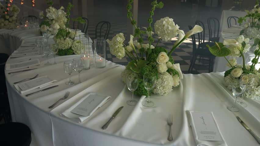 Elegant wedding table setup with lush white floral centerpieces, fine glassware, and formal place settings in a sophisticated reception venue, parallax shot.