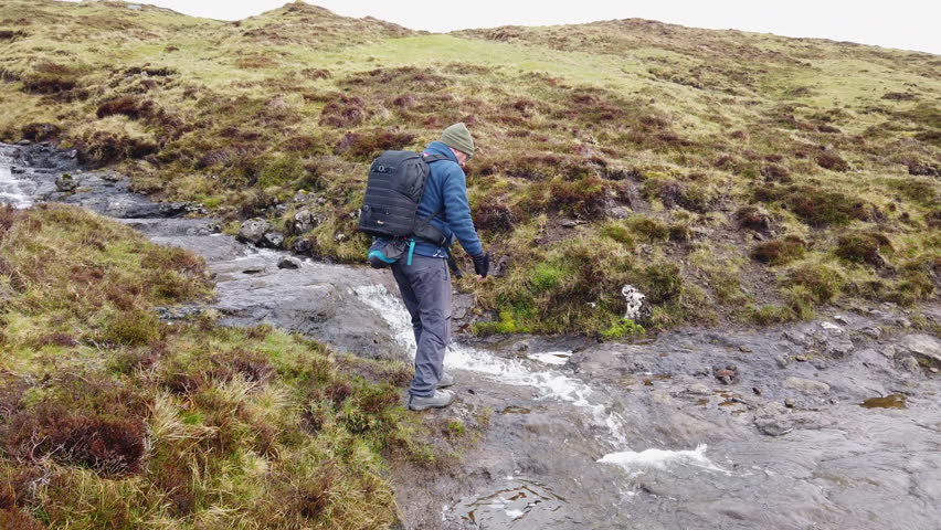Man wearing a backpack and hiking gear jumps over a mountain stream in the Faroe Islands