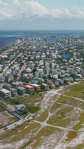 Rows of coastal houses spread across a seaside town as sandy dune pathways weave through grassy terrain, leading toward the shoreline under a summer sky.