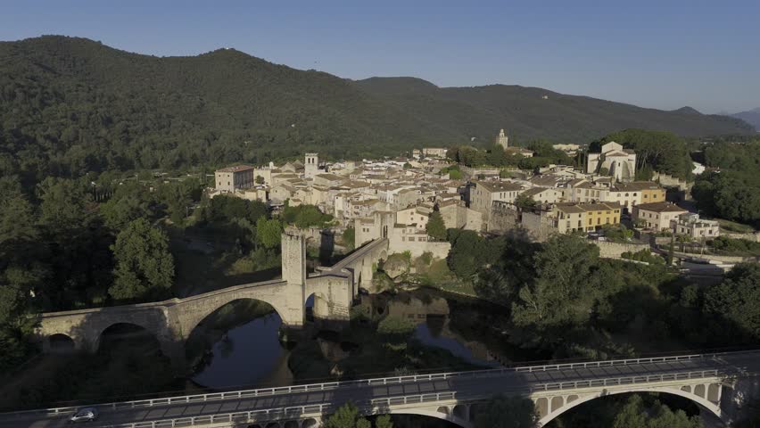Pont de Besalú aerial view with new road bridge in foreground, medieval span over the Fluvià and old town beyond under clear summer light; ideal for travel ads.