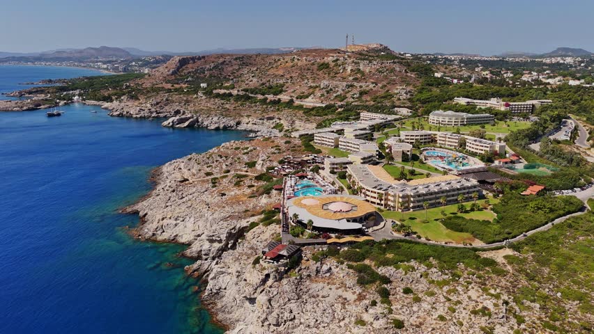 Drone captures the beautiful coastline of Rhodes, Greece, featuring a luxury resort and clear blue waters under a bright sky, perfect for relaxation and exploration.