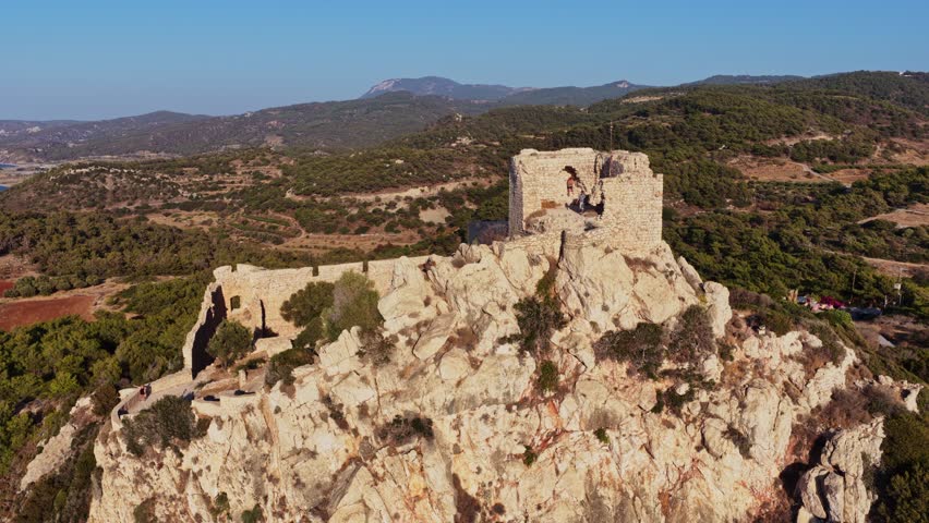 Aerial view of a historic stone fortress sitting atop a rocky hill in Rhodes. Surrounded by lush greenery and distant mountains, the scene captures natural beauty.