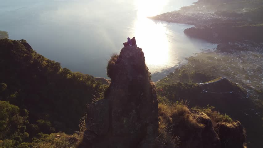 Captivating 4K drone overhead shot of a couple sitting on a rocky peak above Lake Atitlan at sunrise, with golden reflections across the water and lush landscapes below. Great for travel videos, lifes