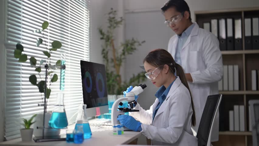 Two scientists are working in a lab, one of them is looking at a computer screen. The lab is filled with various scientific equipment and books