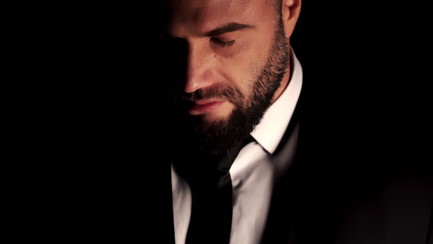 A close-up portrait of a rugged, bearded man in a white shirt and suit against a dark background. A businessman against a dark background looks into the camera.