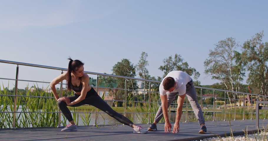 Outdoor view of healthy fit couple doing stretching exercises in nature near river, enjoying fitness routine and healthcare practices in open air, preparing for workout or running
