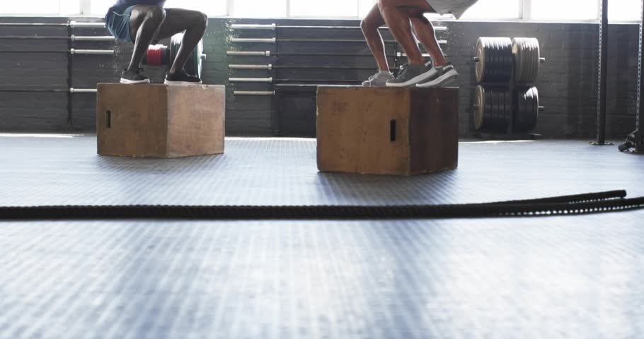 Three men holding squats on plyo boxes and stepping down, initiating synchronized box jumps in gym. Fitness, teamwork, dynamic, athletic, strength, training, motivation