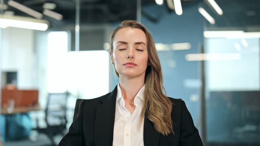 Calm businesswoman in a black blazer finds peace through meditation in a modern office. Her relaxed posture and closed eyes signify stress relief and mental focus for work-life balance.